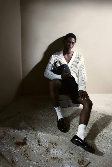 Man in knit cardigan sitting indoors holding dark purse, wearing black shorts and white socks with loafers in soft studio lighting.