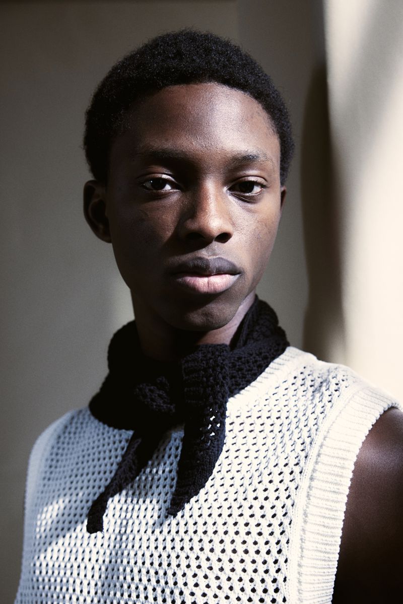 Portrait of a young Black man wearing a white knit tank top and a black knit scarf indoors against a neutral wall.