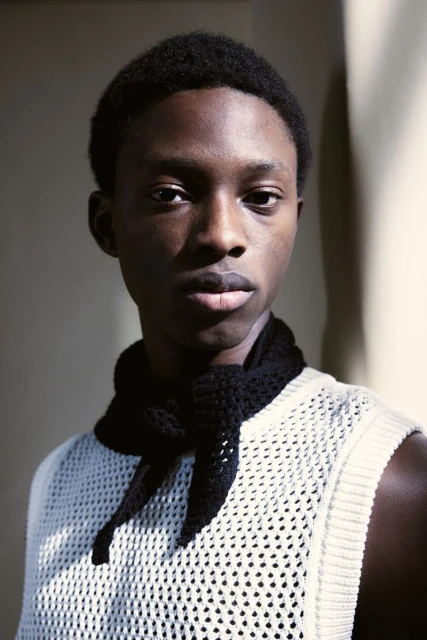 Portrait of a young Black man wearing a white knit tank top and a black knit scarf indoors against a neutral wall.
