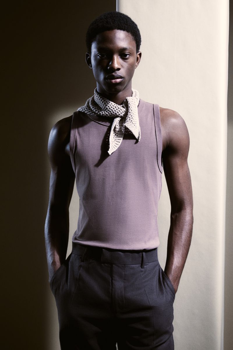 A young adult Black male model stands in a studio wearing a ribbed tank top, dark trousers, and a knit kerchief around his neck.