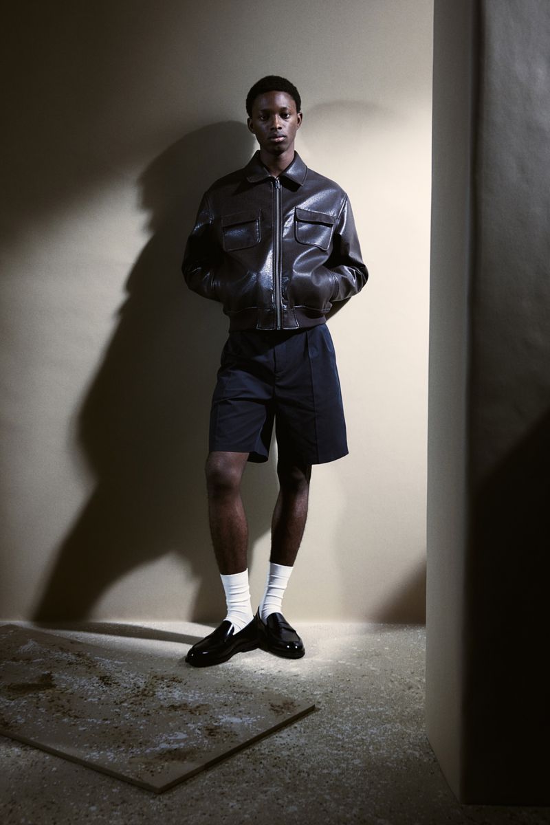 Full-length shot of a young Black man in a brown leather jacket and navy shorts, posing against a beige wall in a dimly lit interior.