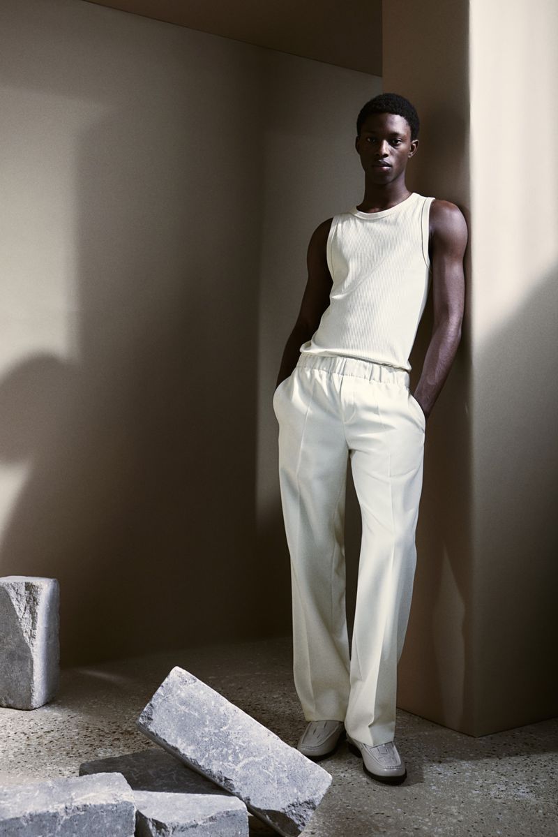 Fashion portrait of a Black male model wearing a tank top and cream pants leaning against a wall in a photo studio with stone blocks.