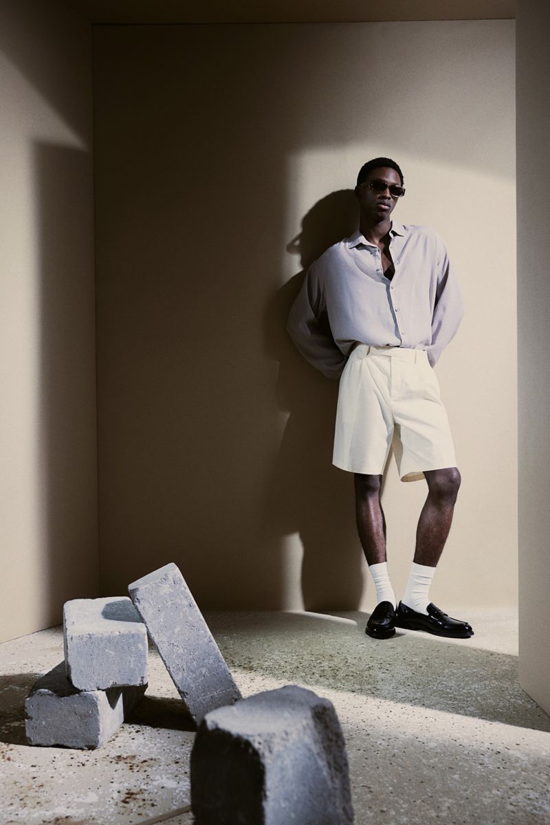 Fashion model poses in a beige setting, wearing a light purple shirt, white shorts, white socks, and black loafers next to concrete blocks.