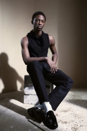 Black man seated on stacked concrete blocks wearing a black tank top and trousers in a studio setting