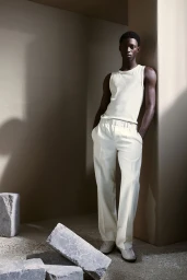 A Black male model wearing an off-white tank top and matching trousers leaning against a concrete pillar in a minimalist interior.