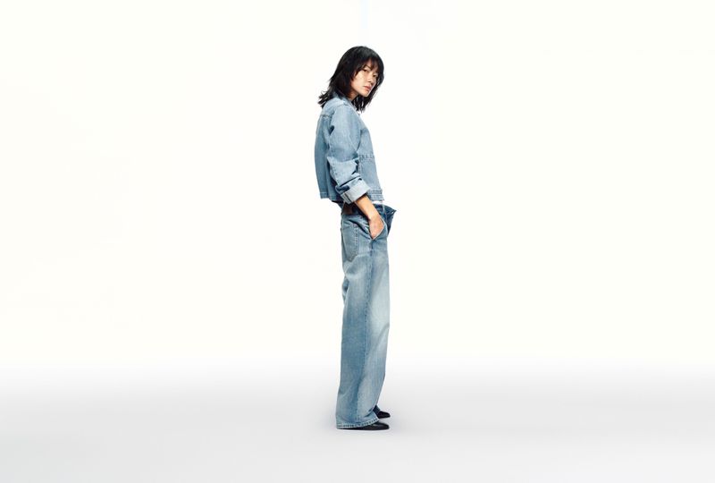 Asian woman poses in light-blue denim jacket and wide-leg jeans against a white backdrop in a fashion studio shot, showcasing material and cut.