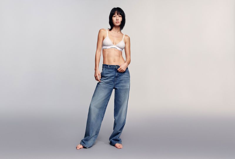 An Asian woman poses in a white bra top and baggy blue jeans in a full shot against a solid light gray studio backdrop.