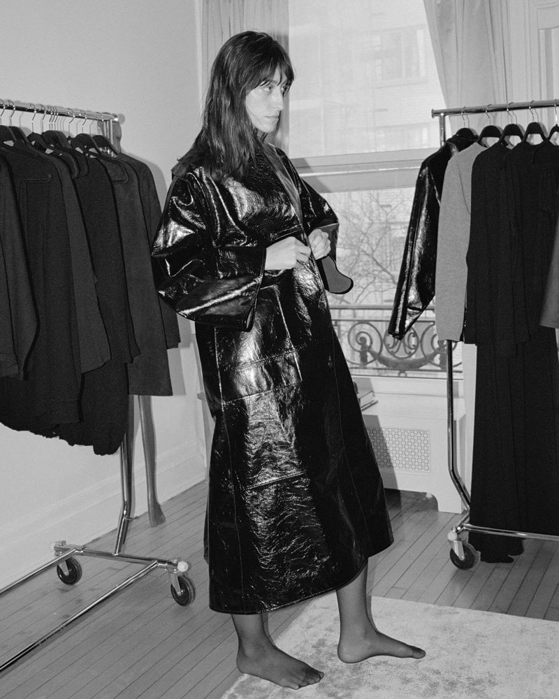A woman poses indoors, wearing a shiny black coat, with clothing racks and a window in the background, in a high-contrast, black-and-white image.