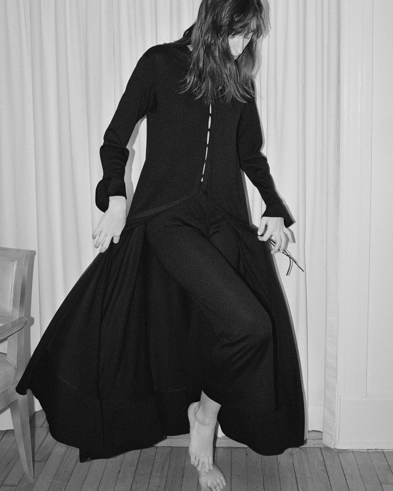 Monochrome image of a woman in a long black dress, standing indoors with bare feet on a wooden floor, against a white draped backdrop.