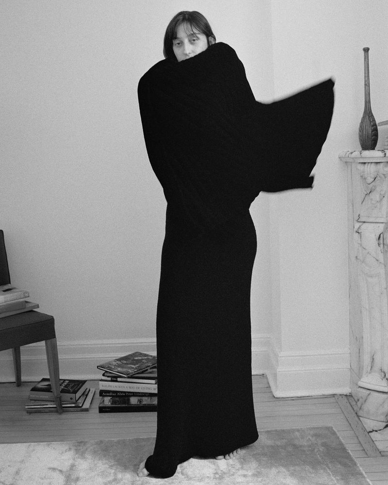 Model wrapped in a heavy black knit garment standing in a light domestic interior with books on the floor.