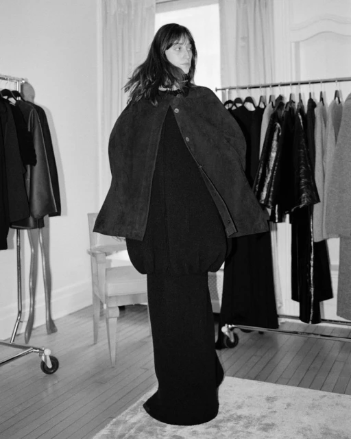 Black-and-white shot of a woman in a black dress and cape standing in a room with clothing racks and a chair.