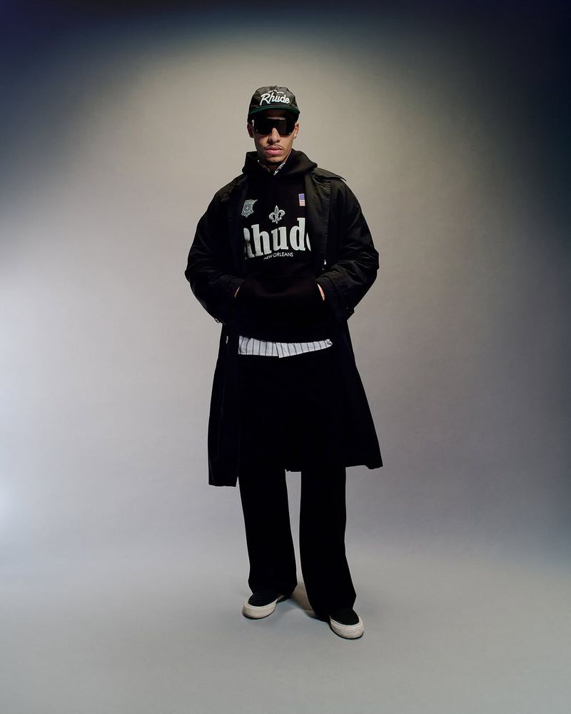 A male model posing indoors in a black hoodie, trench coat, baseball cap, sunglasses, black pants, and sneakers in a studio setting.
