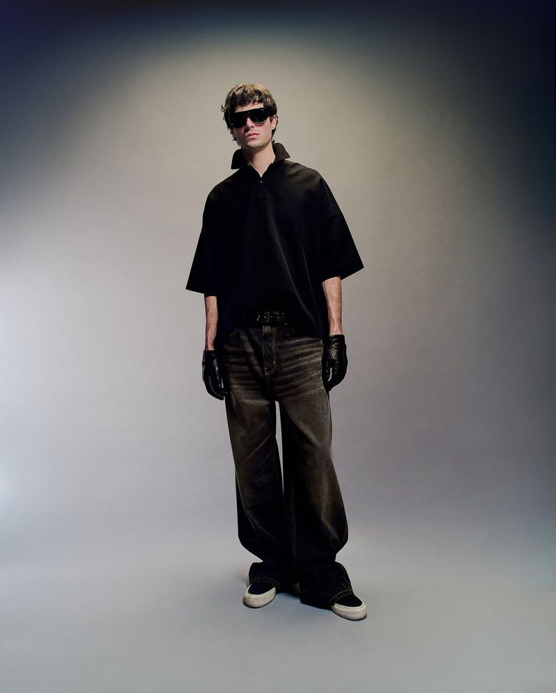 Fashion shot of a young adult male model wearing a black polo shirt, brown jeans, black gloves, and sneakers in a studio setting.