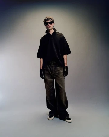 Fashion shot of a young adult male model wearing a black polo shirt, brown jeans, black gloves, and sneakers in a studio setting.