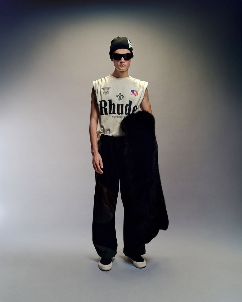 Man in sleeveless shirt, black pants, beanie, and sunglasses stands in a photo studio, holding a black coat, fashion photography.