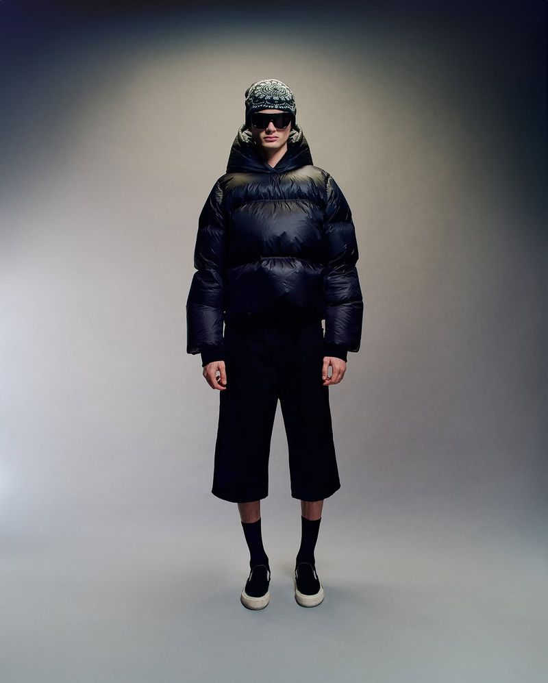 Model wearing a black puffer jacket, sunglasses, and patterned beanie against a neutral grey studio backdrop.