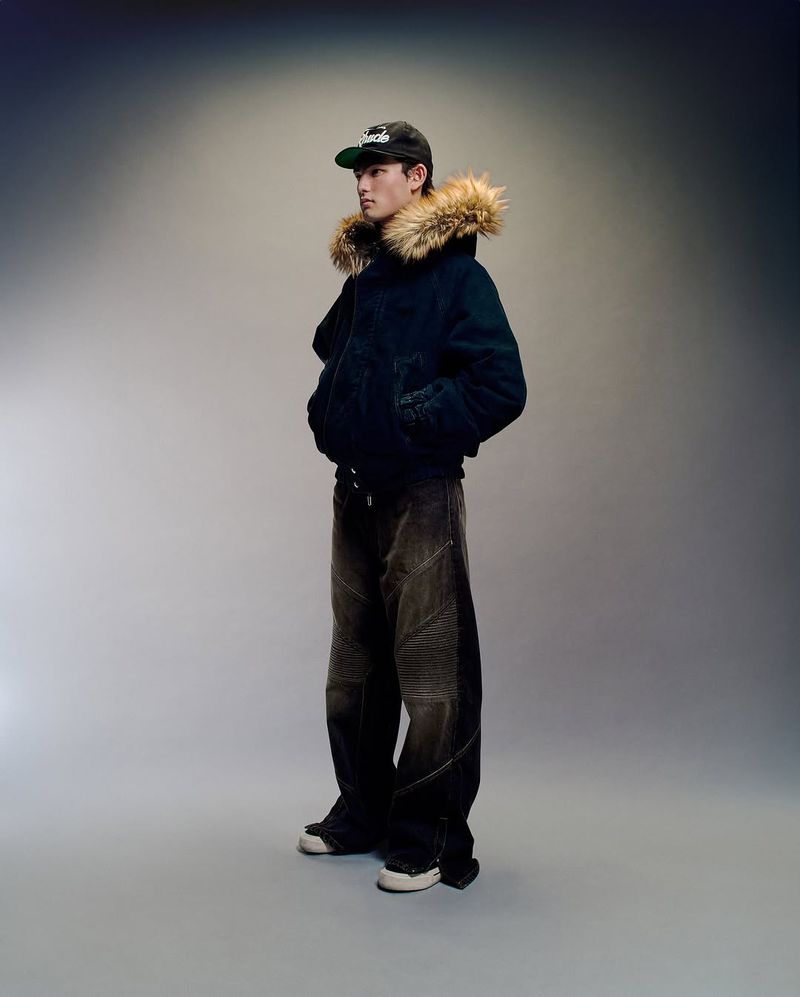 Full body shot of a person in a winter jacket with a fur-lined hood, baseball cap, and dark denim jeans against a grey studio background.