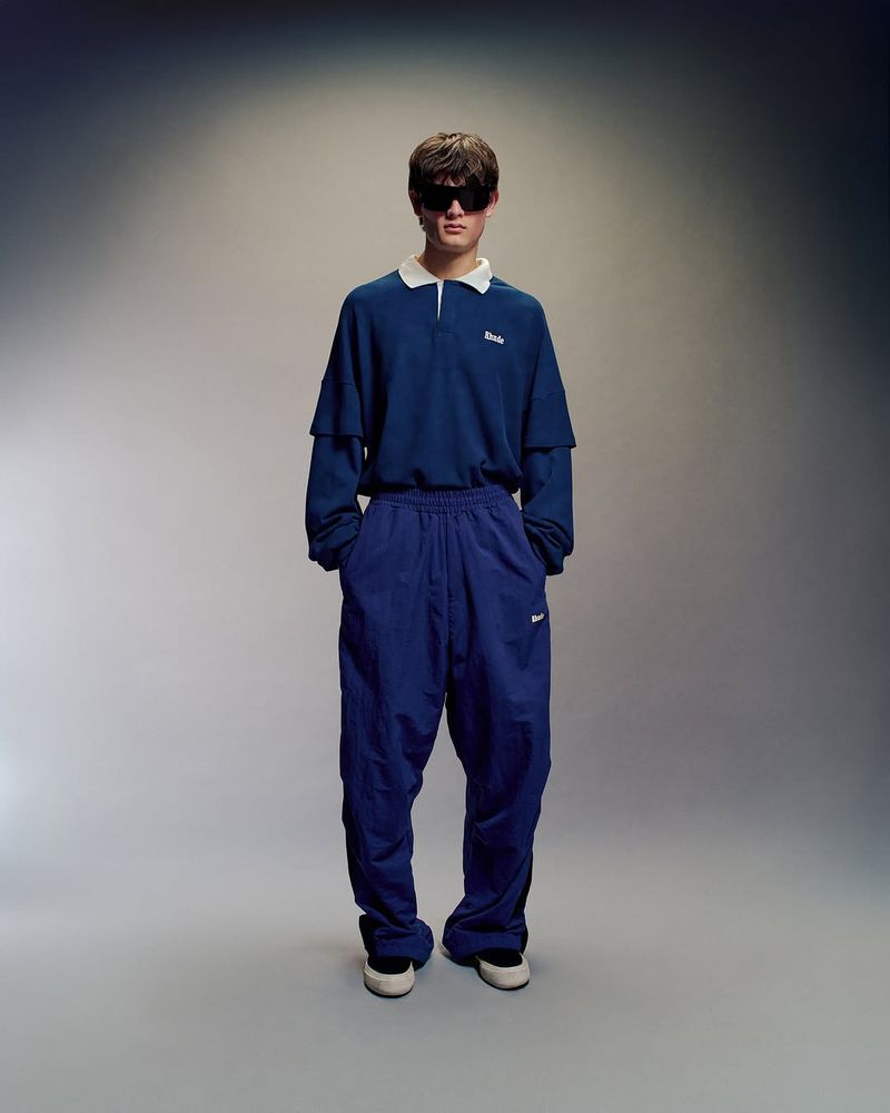 Full-length shot of a young adult male in a blue collared shirt and track pants, posing against a neutral studio backdrop with soft, diffused lighting.