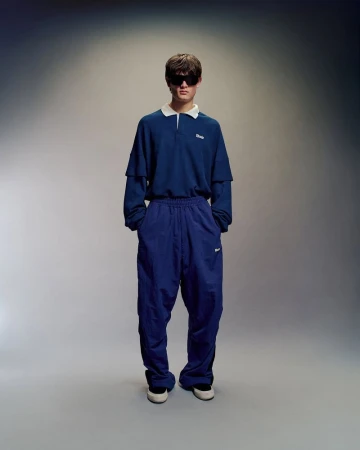 Full-length shot of a young adult male in a blue collared shirt and track pants, posing against a neutral studio backdrop with soft, diffused lighting.