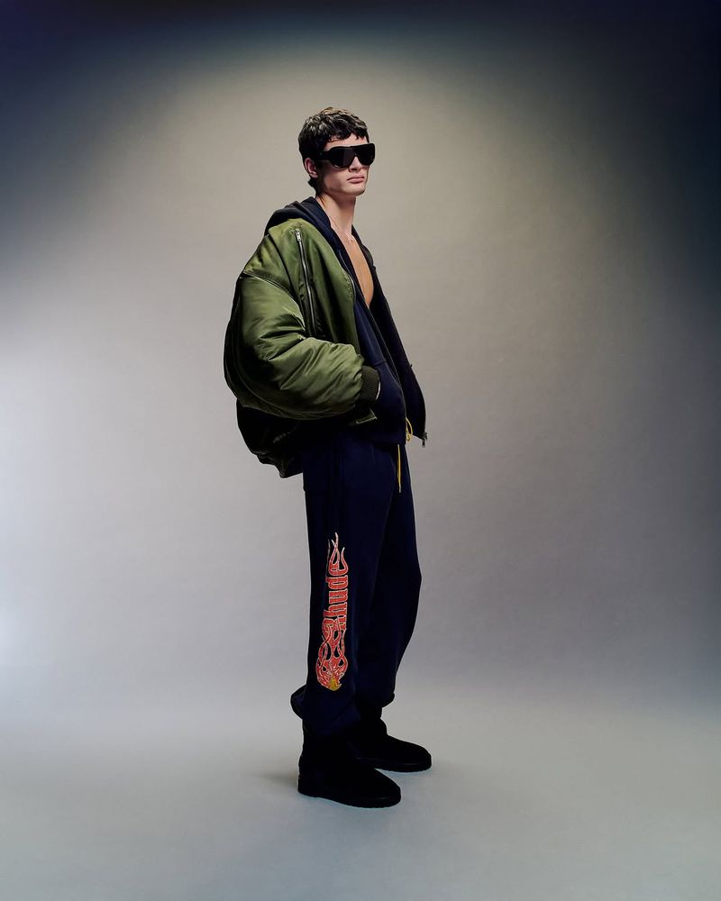 A young man in a bomber jacket and sweatpants stands in a photo studio with neutral gray walls and floor and soft light. He wears sunglasses.