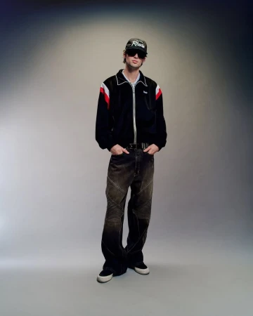 Model wearing a black zip jacket with red and white stripes, dark jeans, and a cap against a grey studio backdrop