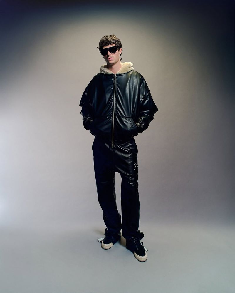 Full-length shot of adult male model wearing a black leather bomber jacket with sunglasses in a photo studio setting.