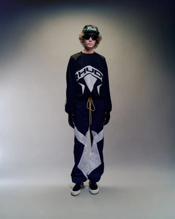 Model in black sweatshirt and dark trousers with white graphic panels standing against a grey studio backdrop.
