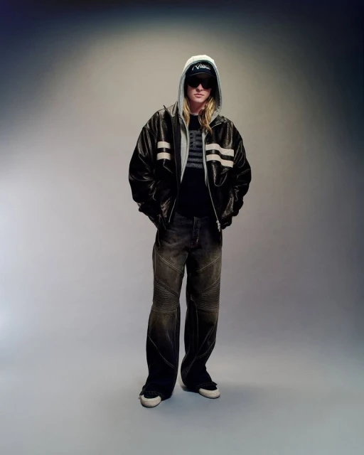 Adult male with blonde hair and sunglasses poses wearing a hooded jacket, t-shirt, baggy jeans, and sneakers in a studio setting.