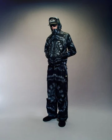 Person in a black and white tie-dye hooded jacket and pants, wearing a black cap and sunglasses, posing in a studio.