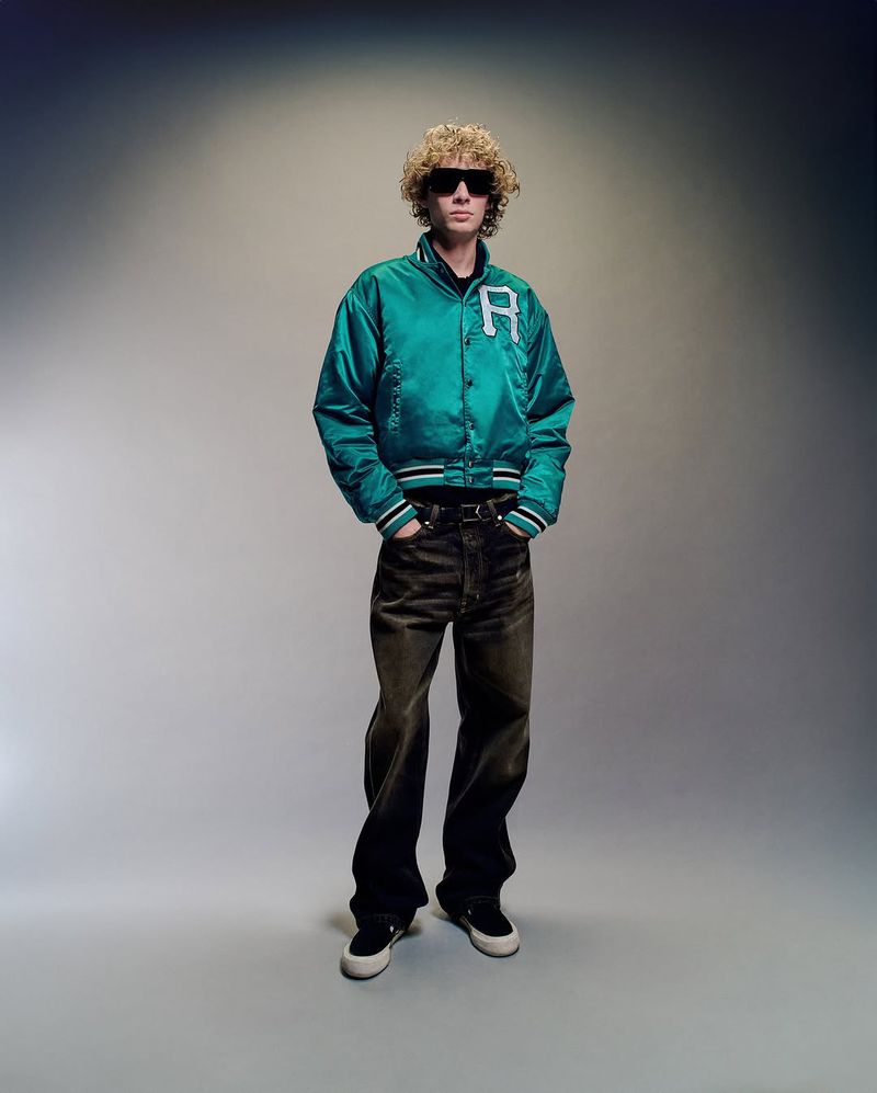 Full-length shot of a young adult with curly hair wearing sunglasses, a letterman jacket, dark pants, and sneakers in a photo studio.