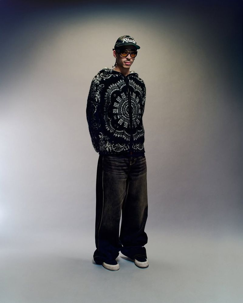 Full-length shot of an adult male wearing a black and white patterned hooded jacket, oversized jeans, sneakers, glasses, and baseball cap.