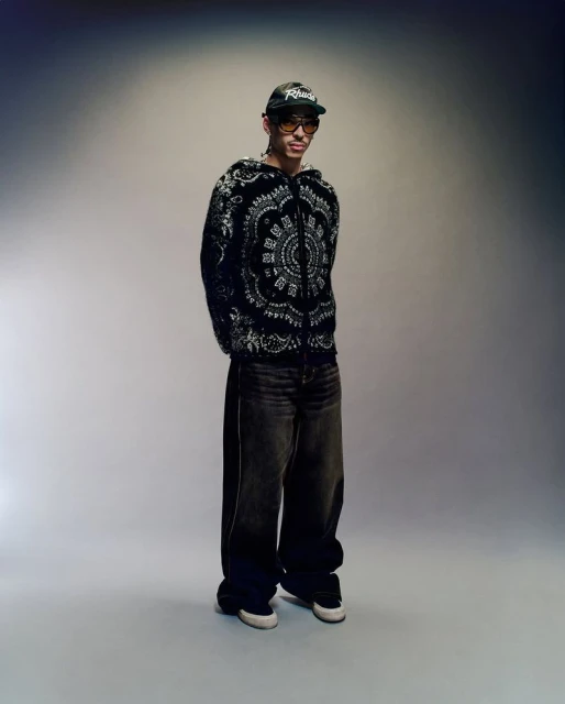 Full-length shot of an adult male wearing a black and white patterned hooded jacket, oversized jeans, sneakers, glasses, and baseball cap.