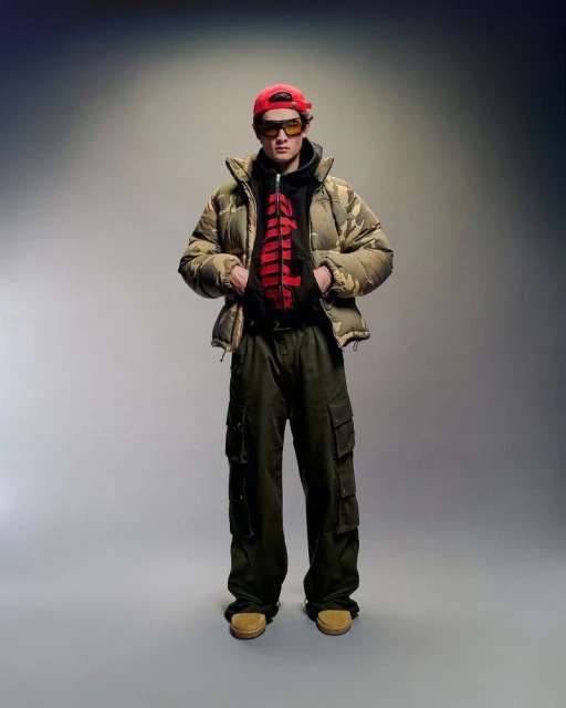 Full-length shot of a male model in a camouflage puffer jacket, red cap, sunglasses, cargo pants, and loafers posing in a studio.