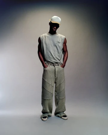 Model wearing a grey muscle tank and trousers standing against a gradient studio background
