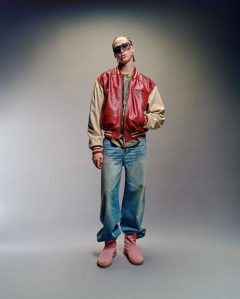 Man in red leather jacket and sunglasses stands against neutral backdrop in full shot, showcasing casual fashion and unique style.