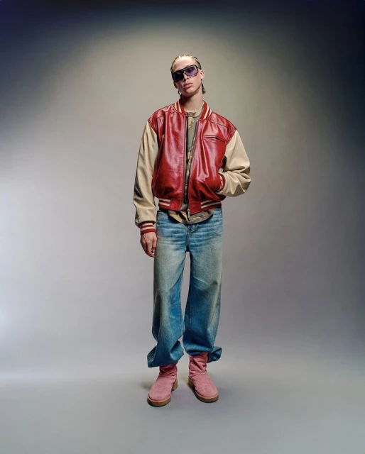Man in red leather jacket and sunglasses stands against neutral backdrop in full shot, showcasing casual fashion and unique style.