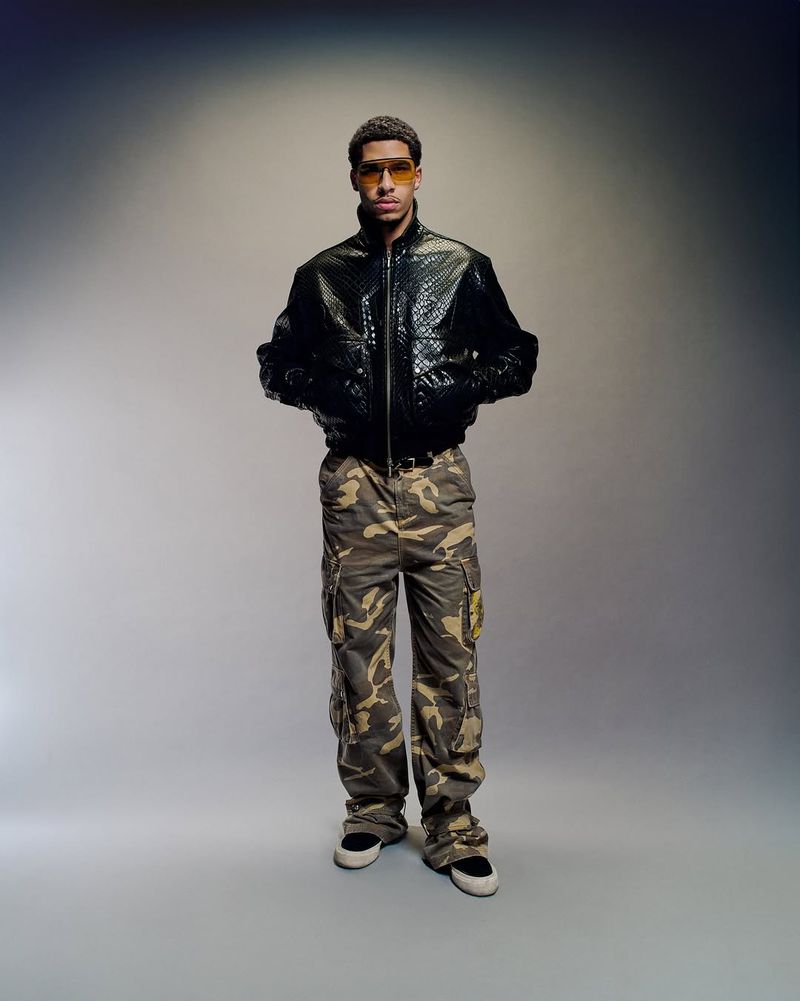 Full shot of a young adult male wearing a black jacket and camouflage pants, posing against a gradient gray background in a studio setting.