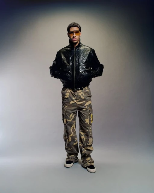Full shot of a young adult male wearing a black jacket and camouflage pants, posing against a gradient gray background in a studio setting.