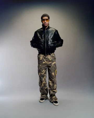 Full shot of a young adult male wearing a black jacket and camouflage pants, posing against a gradient gray background in a studio setting.