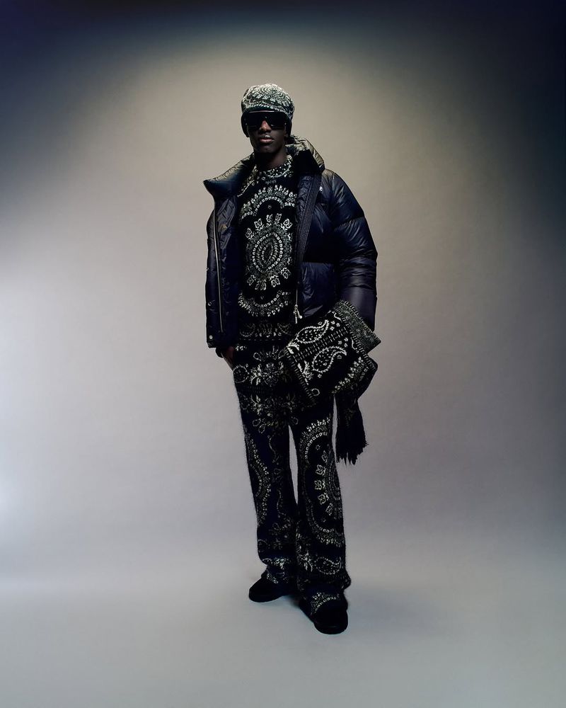 Full shot of an adult male wearing a patterned outfit, jacket, sunglasses, and cap, posing indoors against a neutral background. Fashion.