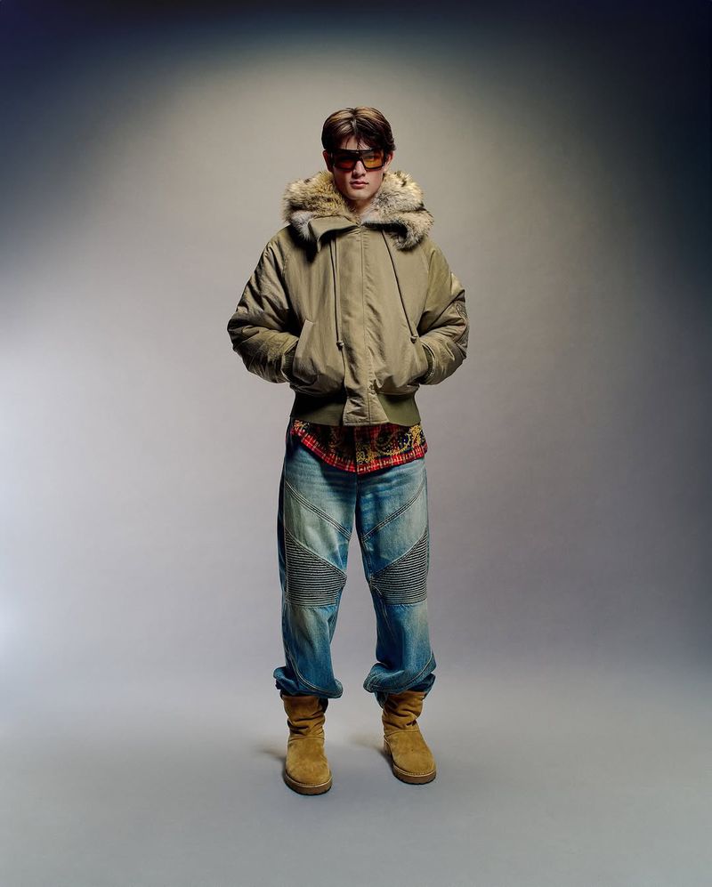 Full-length portrait of a male in a fur-trimmed jacket, jeans, and boots, standing against a gradient grey background in a photo studio.