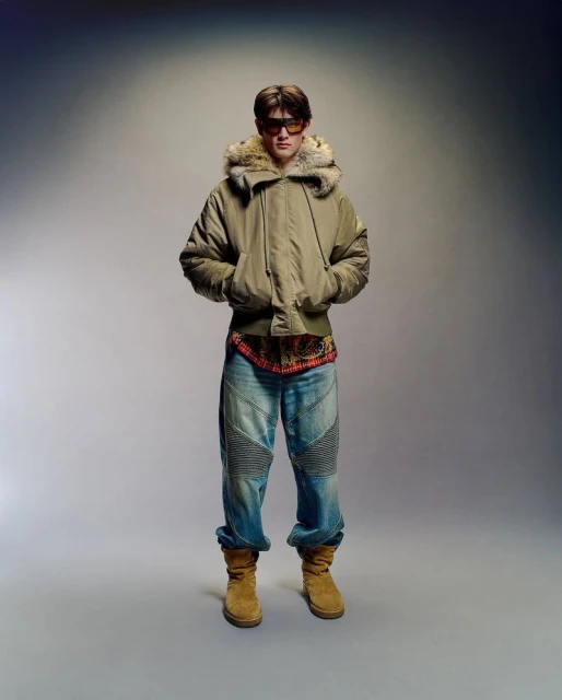Full-length portrait of a male in a fur-trimmed jacket, jeans, and boots, standing against a gradient grey background in a photo studio.