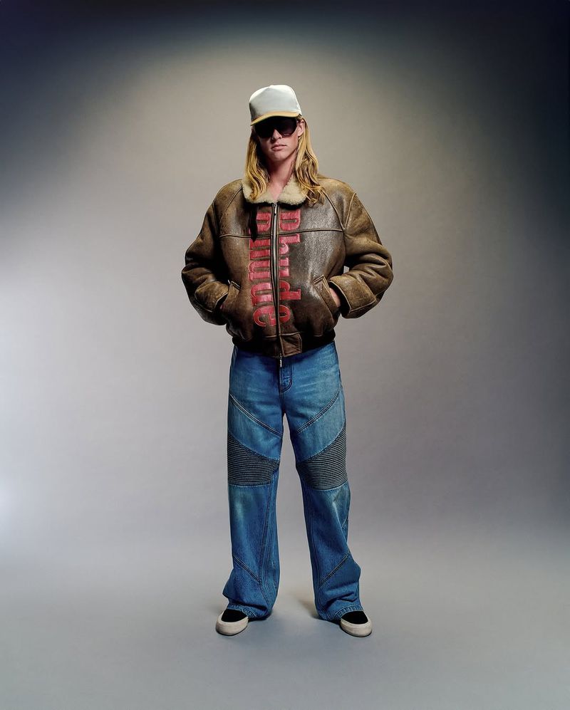 Full-length shot of an adult wearing a baseball cap, sunglasses, shearling-lined jacket, wide-leg jeans, and sneakers in a studio setting.