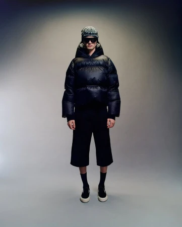 Model wearing a black puffer jacket, sunglasses, and patterned beanie against a neutral grey studio backdrop.