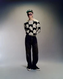 Model standing against a grey gradient backdrop wearing a black and white patterned sweater, dark trousers, and cap.