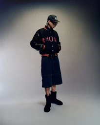 Model wearing a black varsity jacket, dark shorts, cap and boots against a gradient background.