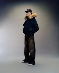 Full-length portrait of a male model wearing a dark winter puffer jacket with fur hood and a baseball cap against a grey backdrop
