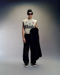 Model wearing dark sunglasses, graphic tank top, and dark trousers holding a textured black coat against a grey gradient studio background
