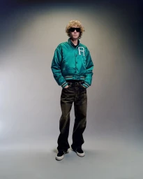 Model with curly blonde hair wearing sunglasses and a green satin bomber jacket in a studio