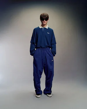 Model wearing dark sunglasses and a blue long sleeve polo shirt with matching blue trousers against a grey backdrop
